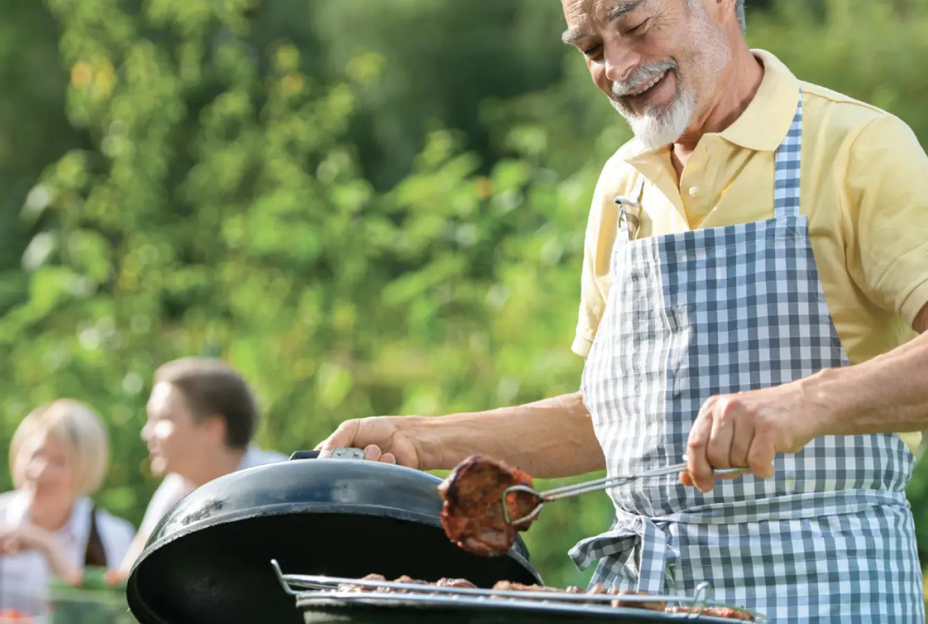 Smiling man with apron holding up a barbeque lid in right hand and tongs picking up the next selection for the grill in the left. A nice sunny day, some guests sitting at a picnic table blurred out in the background.